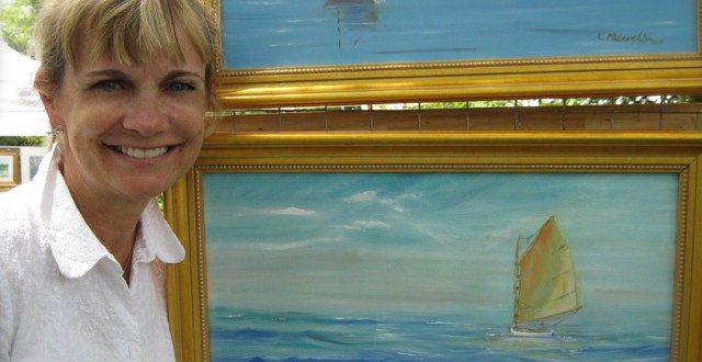 A woman smiles in front of two paintings of sailboats.