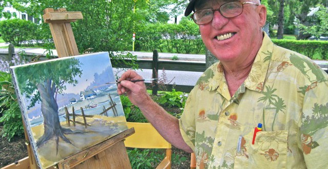 A man in a hat smiles as he holds a painting easel.
