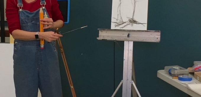 A woman in overalls standing next to an easel.