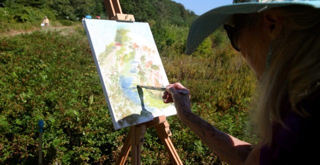 A woman in a hat painting on an easel in a field.
