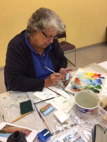 An older woman painting with watercolors at a table.