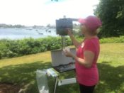 A woman in a pink shirt painting on an easel in front of a boat.