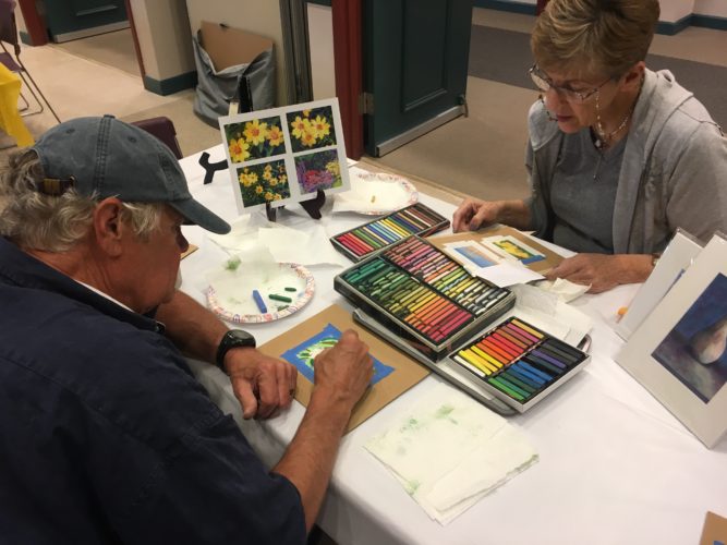 Two people sitting at a table with crayons and paint.