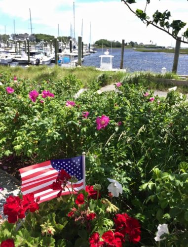 A red, white, and blue American flag waving proudly in a garden near a marina, showcasing the patriotic spirit of The Guild of Harwich Artists.