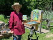 A woman in a hat standing next to an easel in a park.