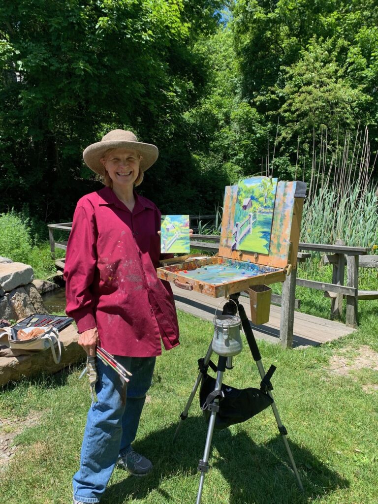 A woman in a hat standing next to an easel in a park.