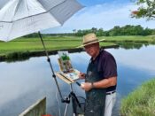 A man holding an umbrella while painting near a body of water.