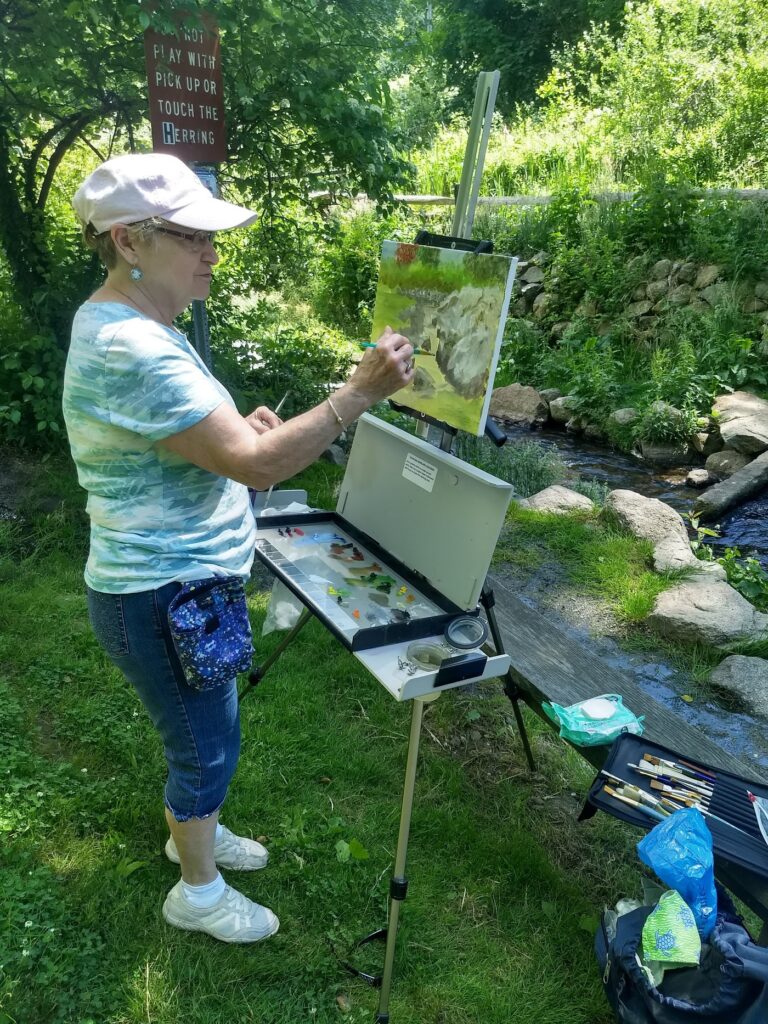 A woman is painting on an easel near a stream.
