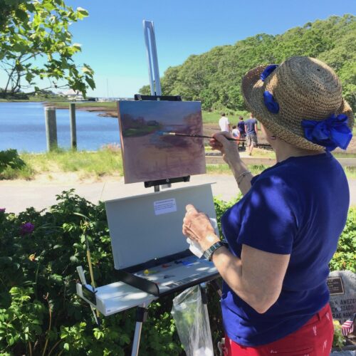 A woman, a member of the Guild of Harwich Artists, painting on an easel near a body of water.