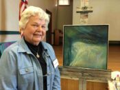 An older woman, representing The Guild of Harwich Artists, stands proudly in front of a painting.